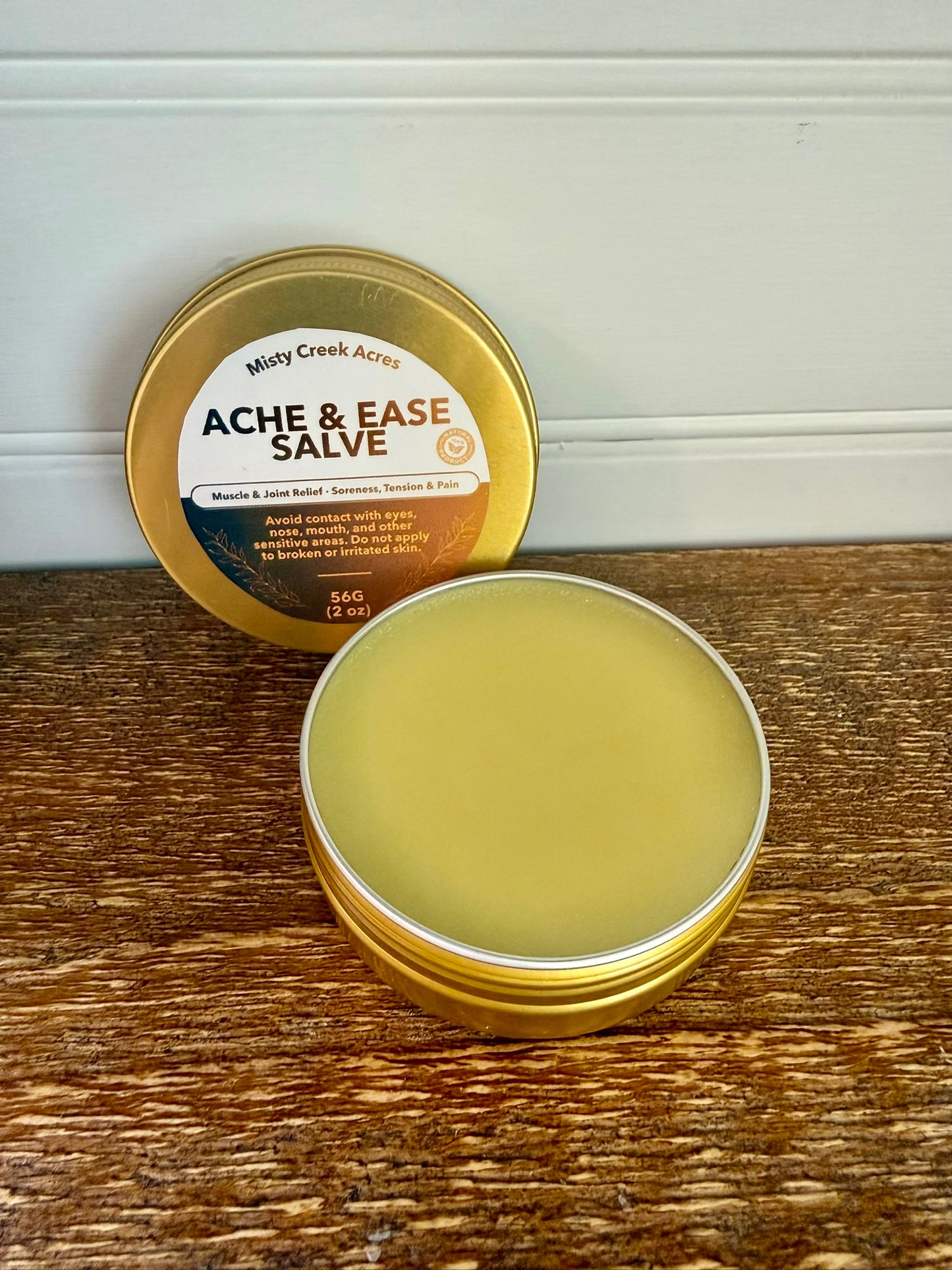 Metal tin of Ache & Ease Salve with a lid on a wooden surface