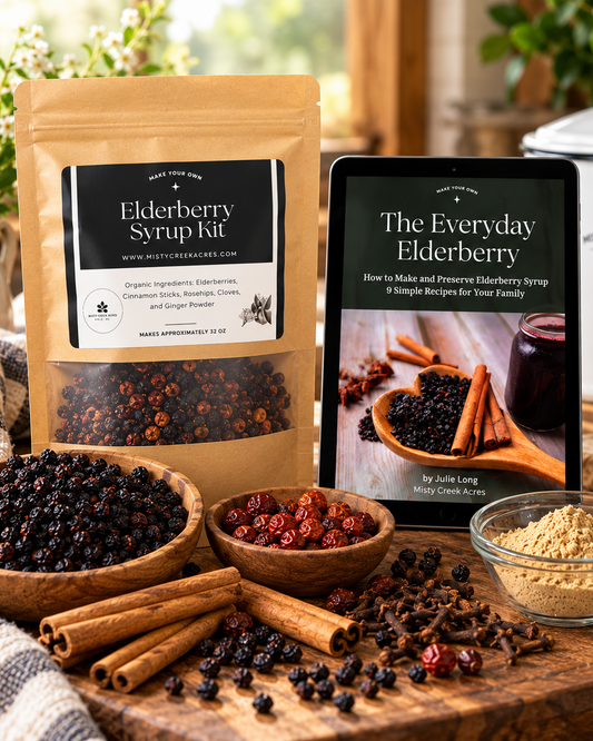 Elderberry syrup kit products displayed on a rustic wooden table: a stand-up pouch labeled Elderberry Syrup Kit, a tablet showing 'The Everyday Elderberry' book, bowls of whole berries, dried berries, cinnamon sticks, and sugar powder, with a kitchen setting in the background.