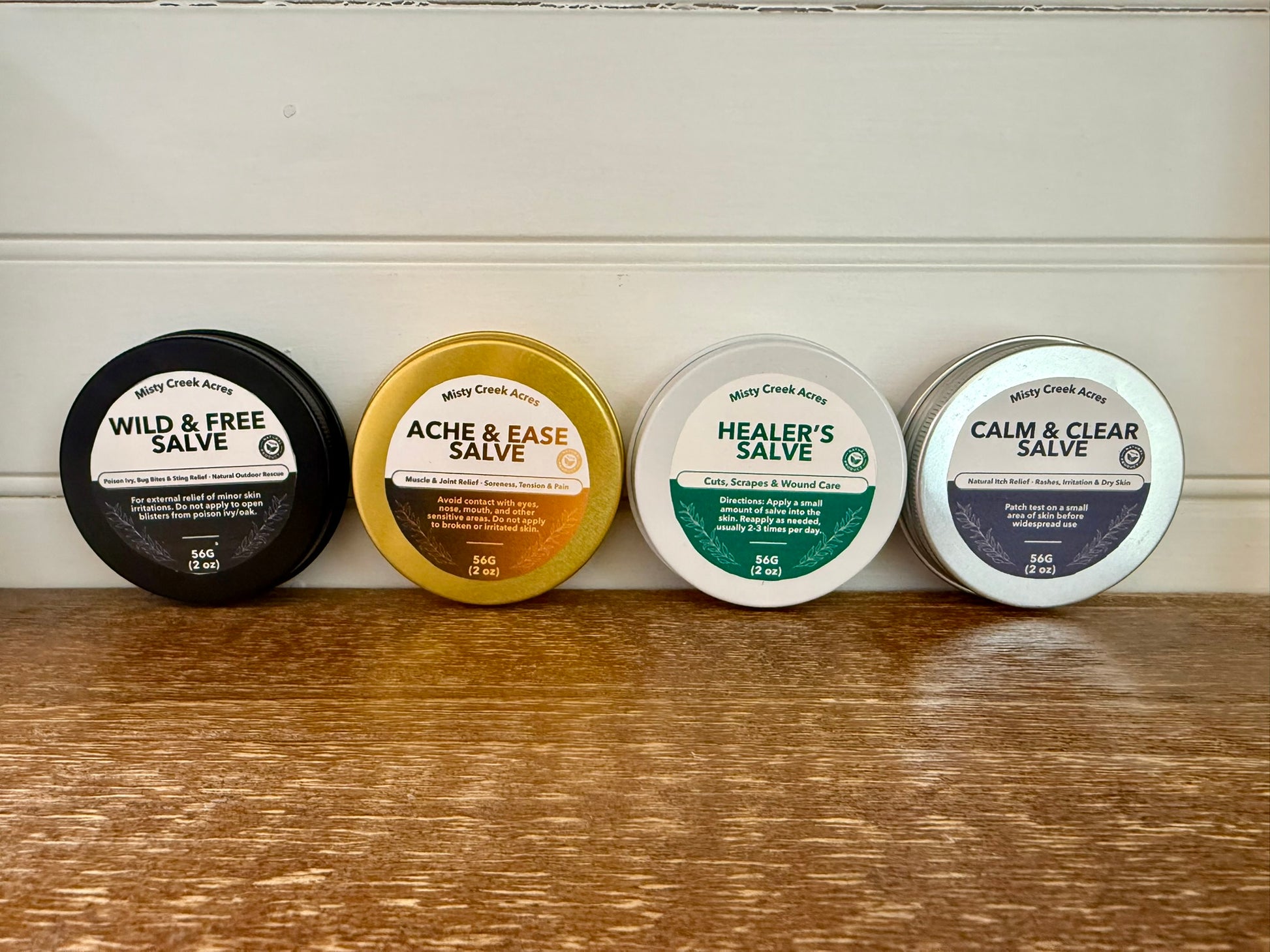 Four tins of Mary Creek Acres salves on a wooden surface with a white background