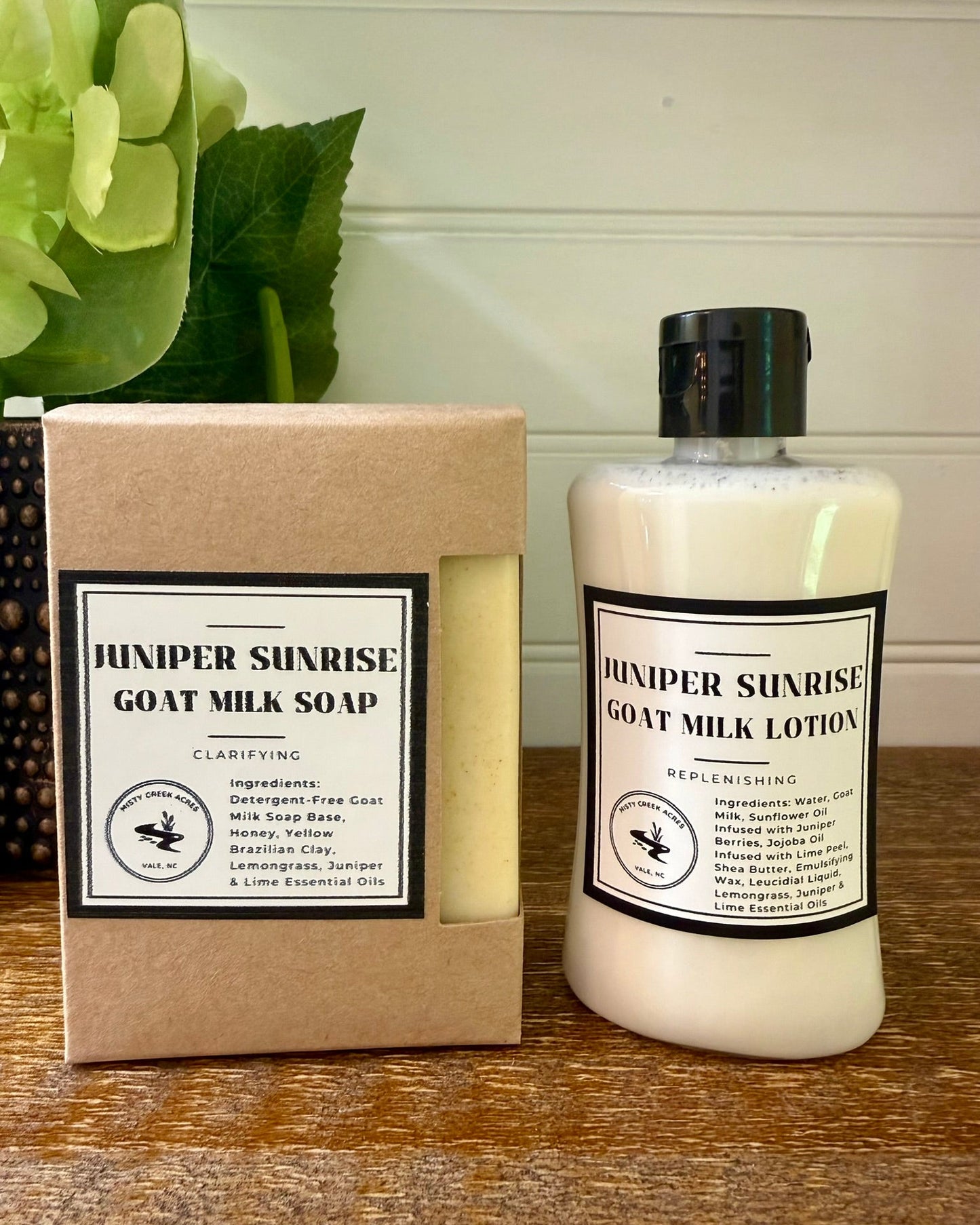 Juniper Sunrise Goat Milk Soap and Goat Milk Lotion set, featuring a kraft paper box of soap and a white bottle of lotion on a wooden surface with a green plant in the background.