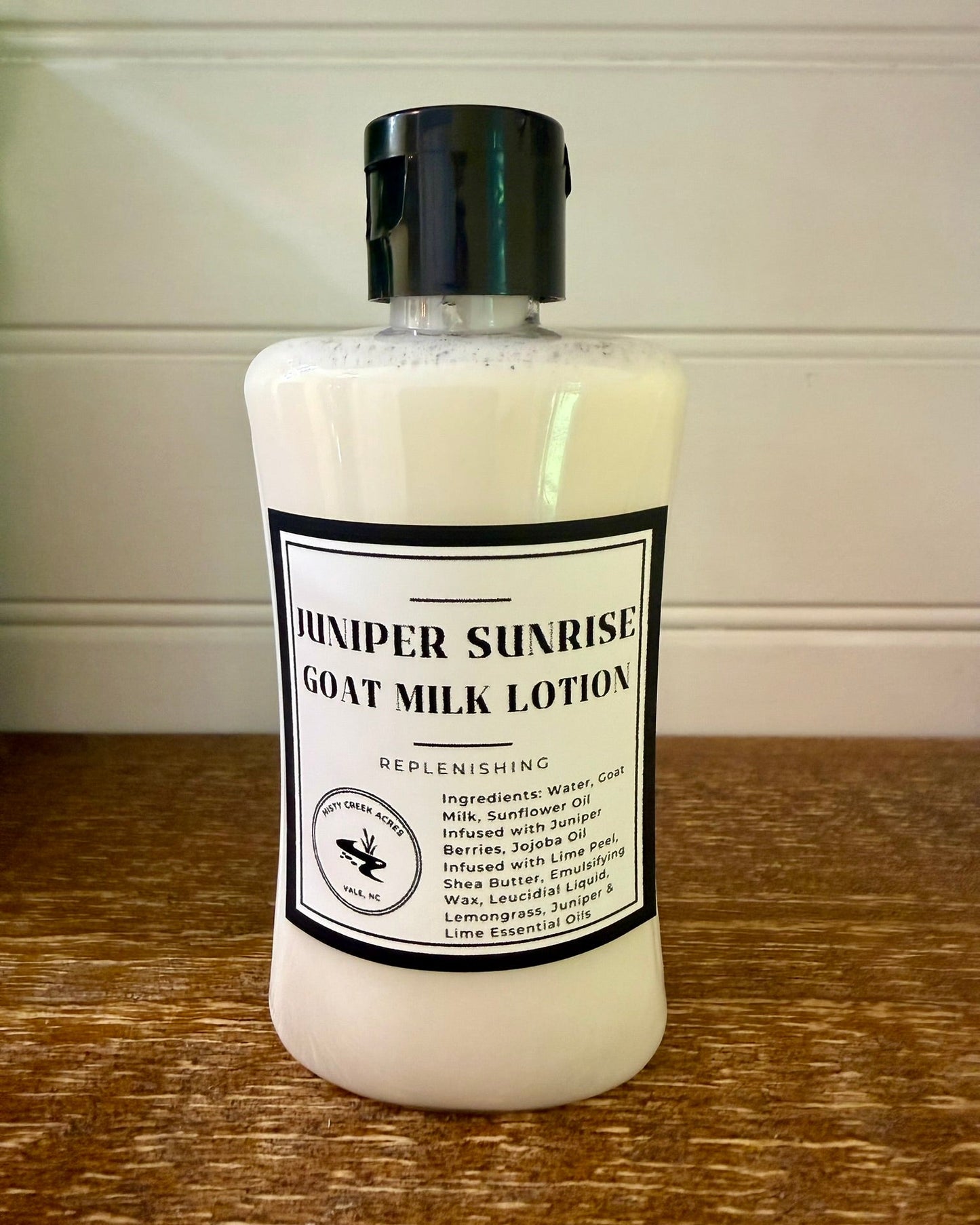 A bottle of Juniper Sunrise Goat Milk Lotion by Wild Balm Co. The lotion is in a white plastic bottle with a black flip-top cap and a black-and-white label, sitting on a wooden surface against a white paneled wall.