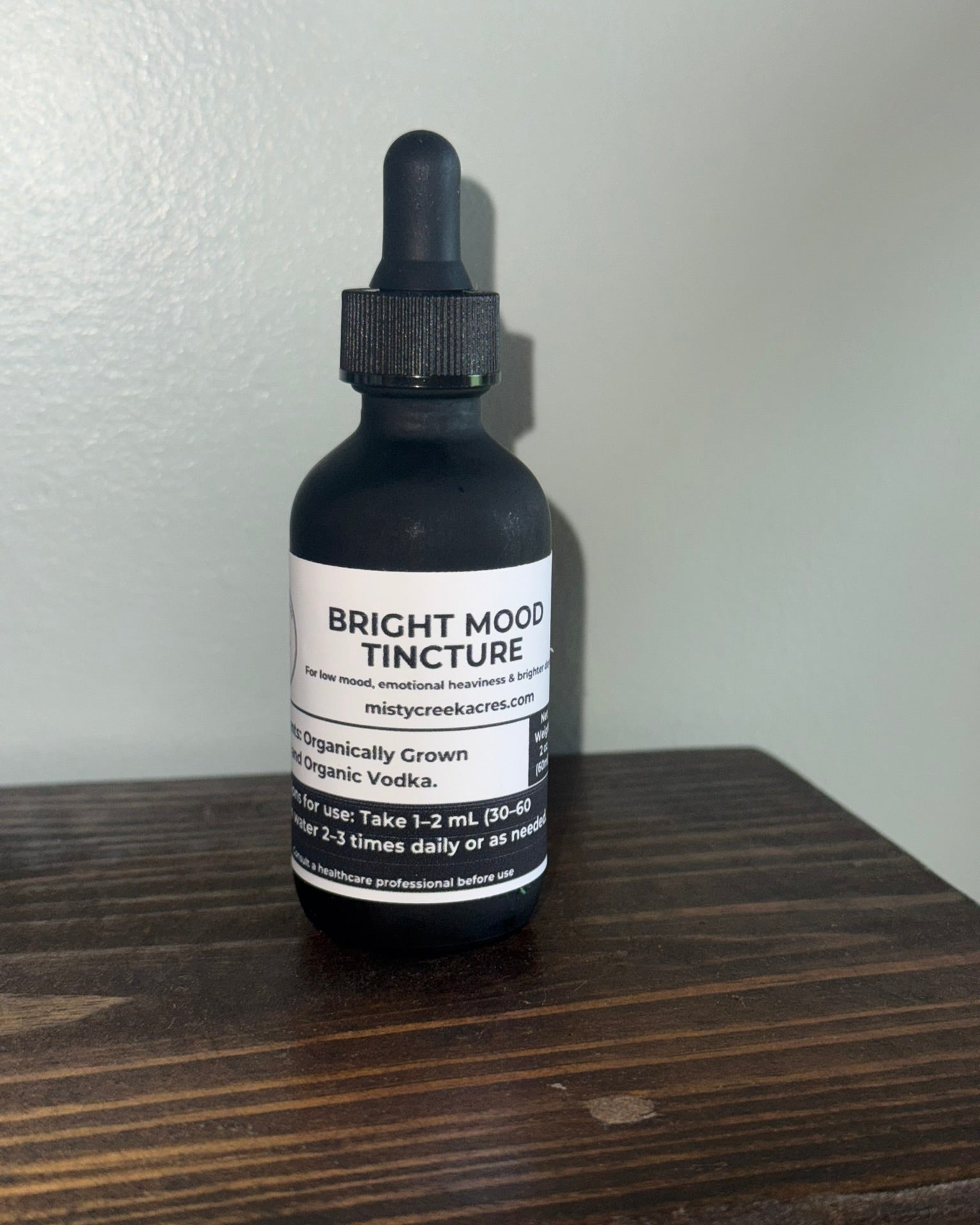 A black glass dropper bottle of Bright Mood Tincture with a white label, sitting on a dark wooden surface against a light gray wall.