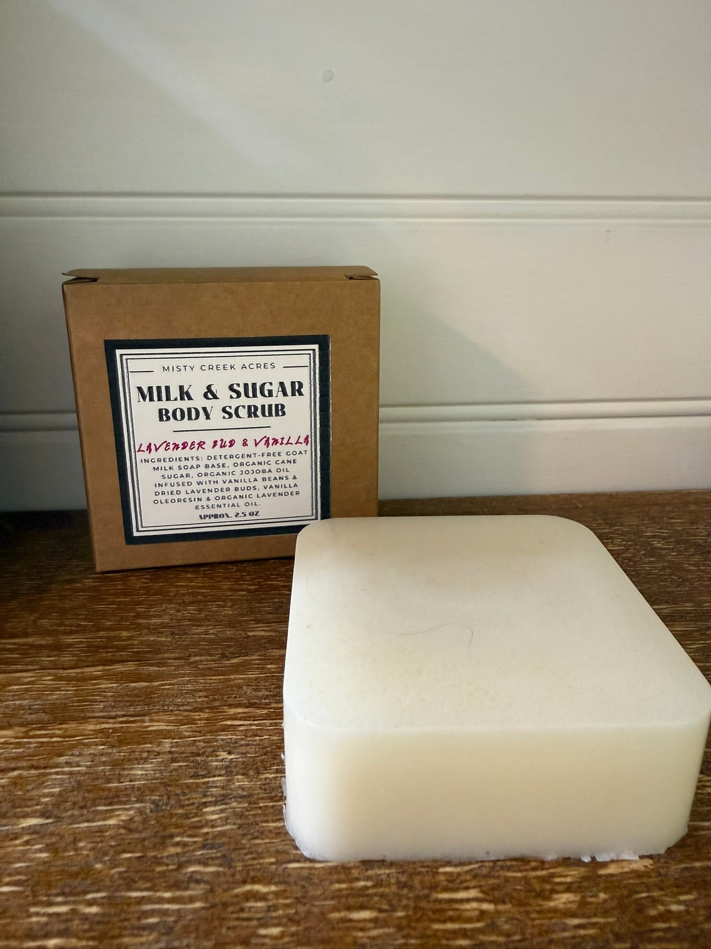 A rectangular bar of off-white body scrub sits on a wooden surface in front of its brown cardboard box, which has a white label reading 'Misty Creek Acres Milk & Sugar Body Scrub'.