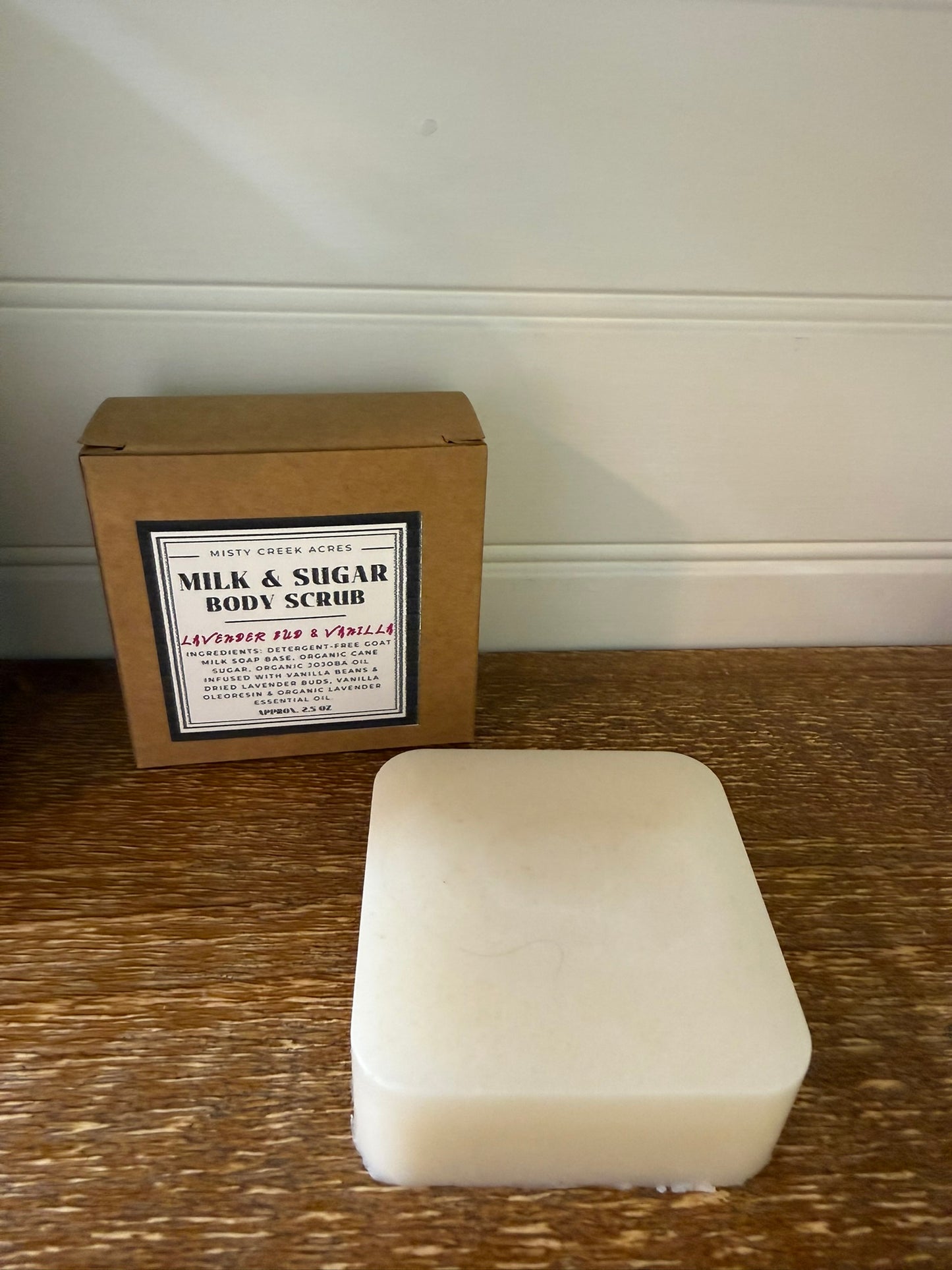 A bar of off-white milk and sugar body scrub sits on a wooden table in front of its brown cardboard packaging box, which features a white label.
