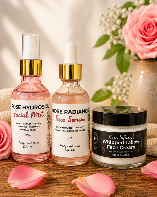 Three skincare products from Misty Creek Acres are arranged on a rustic wooden surface with pink rose petals and a pink rose in the background. The products are: Rose Hydrosol Facial Mist, Rose Radiance Face Serum, and Rose Infused Whipped Tallow Face Cream.