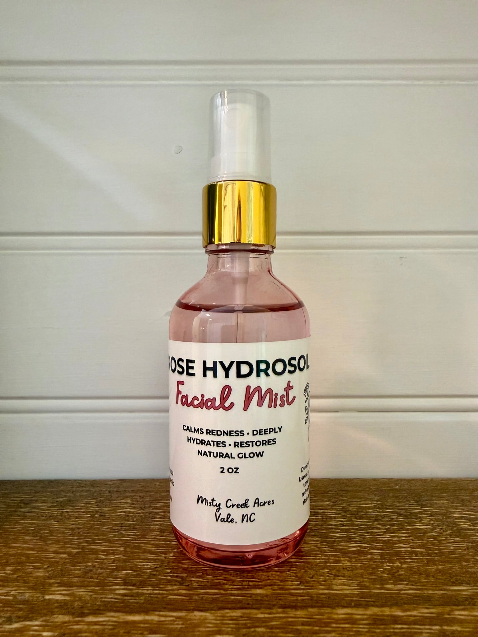 A 2 oz bottle of Rose Hydrosol Facial Mist with pink liquid, a white label, and a gold collar, sitting on a wooden surface against a white paneled wall.