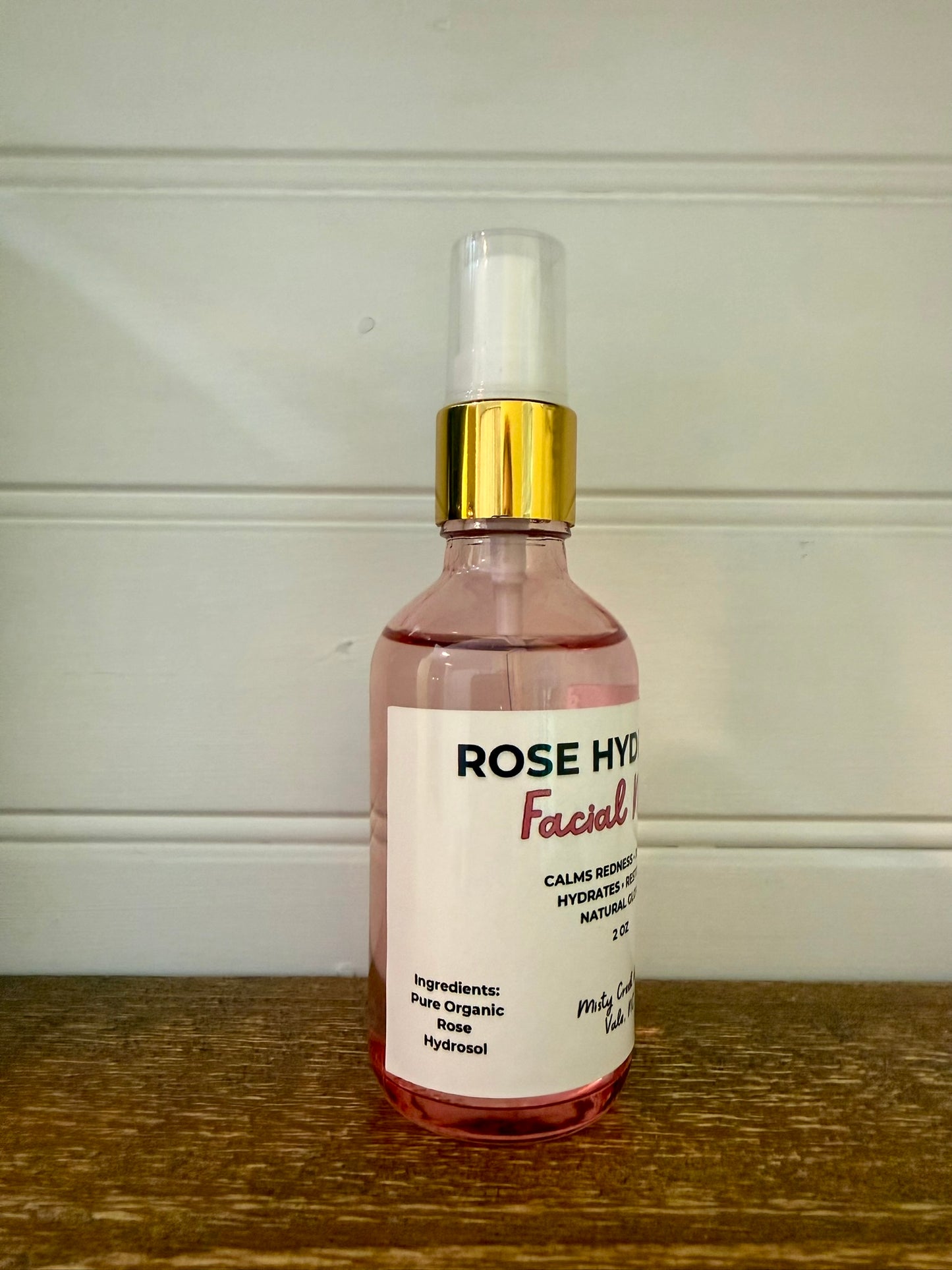 A bottle of Rose Hydrated Facial Toner with pink liquid, a white label, and a gold collar, sitting on a wooden surface against a white paneled wall.