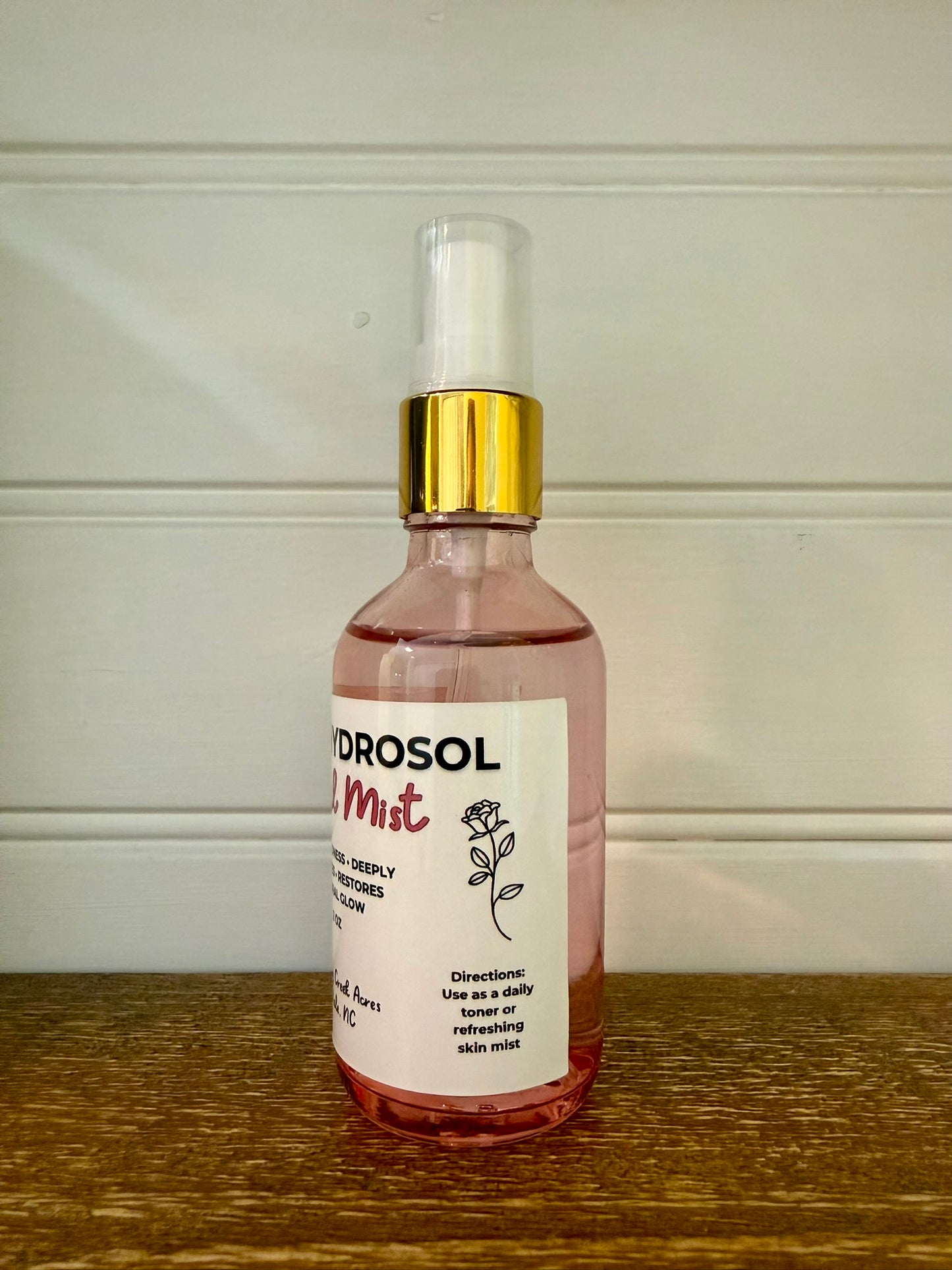 A bottle of Hydrosol Facial Mist with pink liquid, a white label, and a gold collar on a spray pump, sitting on a wooden surface against a white paneled wall.