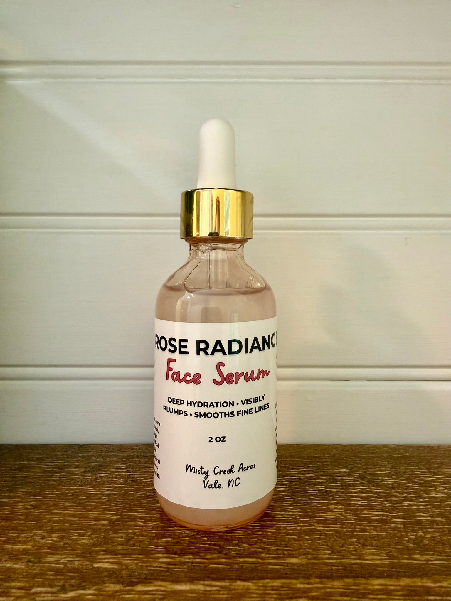 A 2 oz glass dropper bottle of Rose Radiance Face Serum with a white label and gold collar, sitting on a wooden surface against a white paneled wall.
