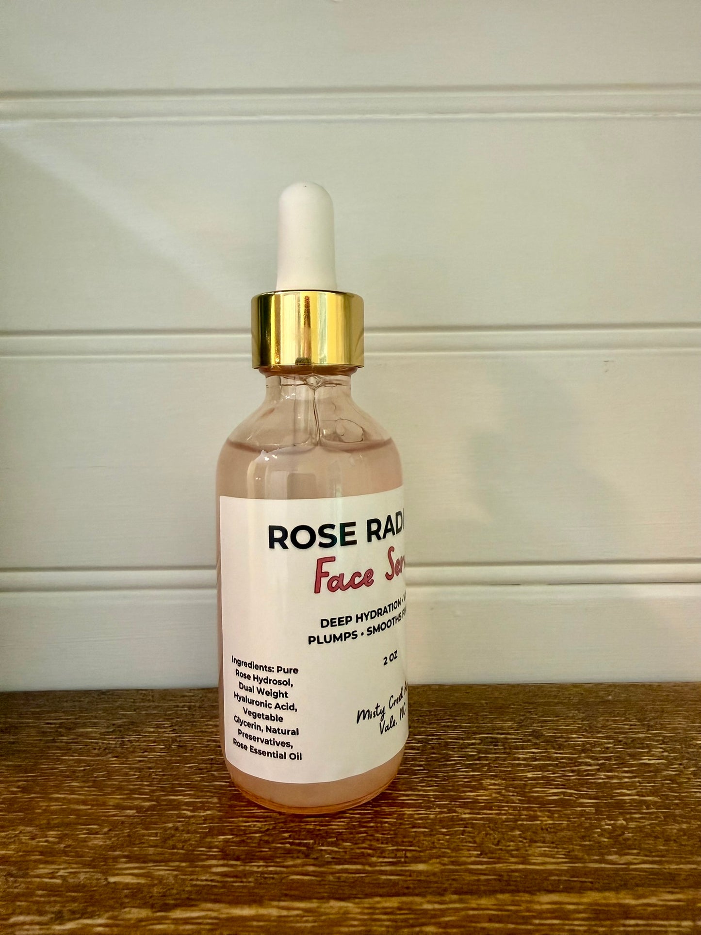 A glass dropper bottle of Rose Rad Face Serum with rose-colored liquid, a white label, and a gold collar with a white dropper cap, sitting on a wooden surface against a white paneled wall.