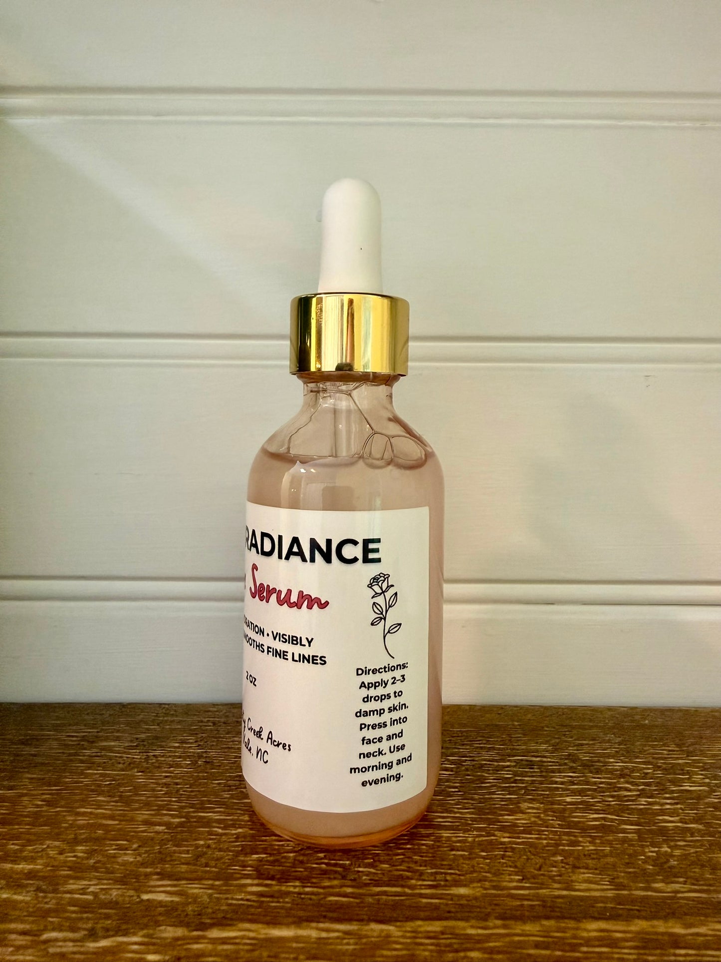 A glass dropper bottle of Radiance Serum with a white label and gold collar, sitting on a wooden surface against a white paneled wall.