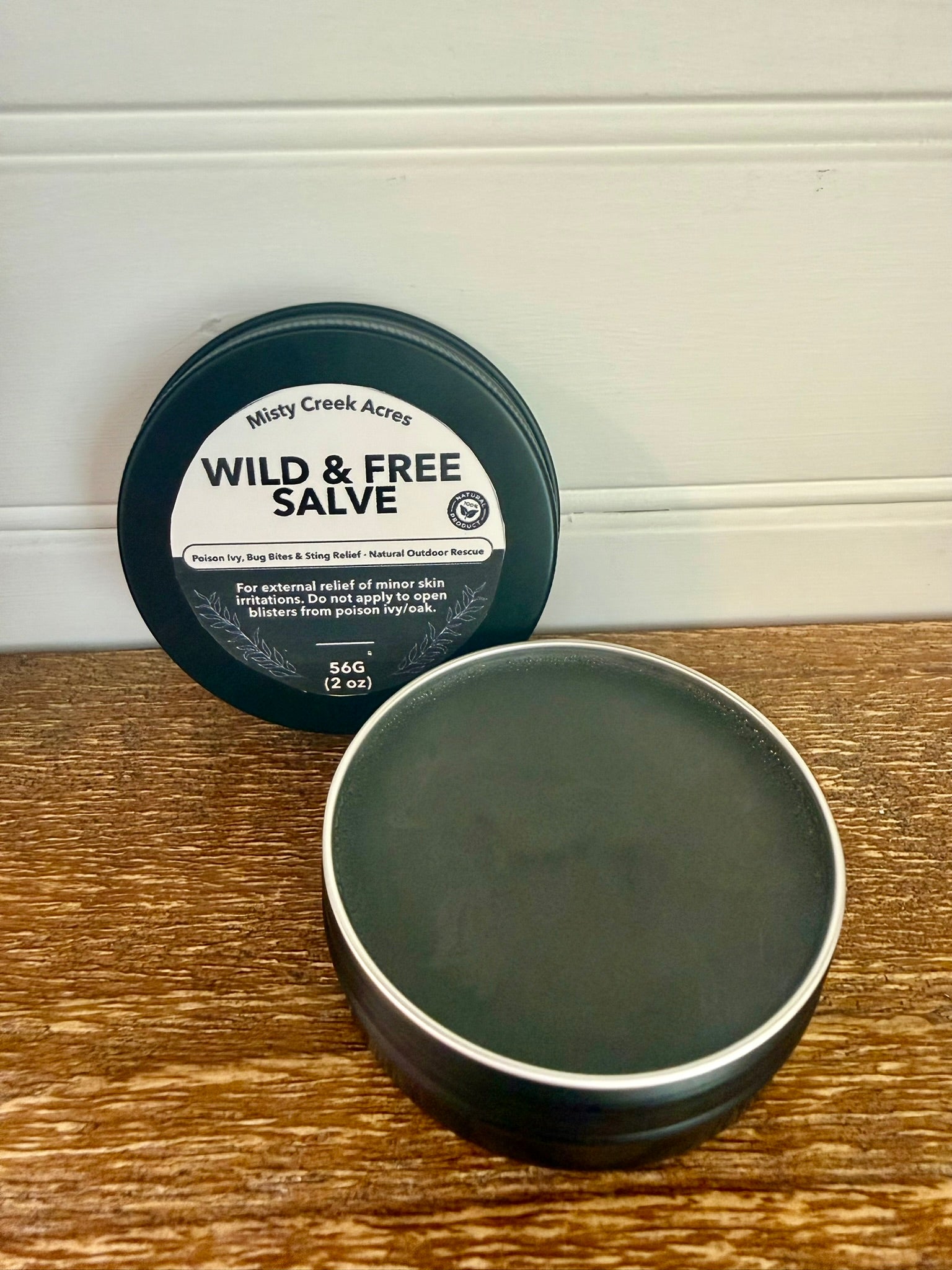 Black jar of 'Wild & Free Salve' by Misty Creek Acres on a wooden surface with a white wall background.