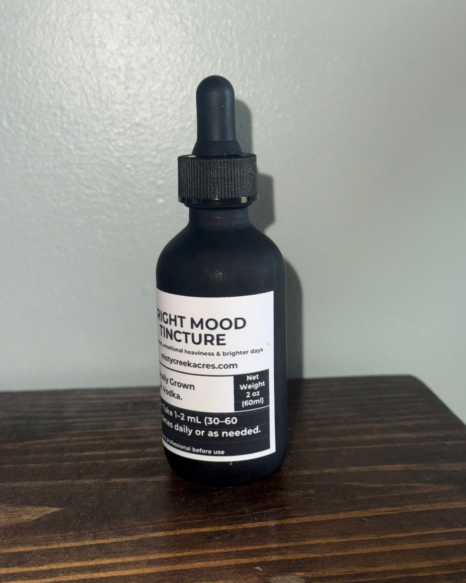 A black glass dropper bottle of 'Light Mood Infusion' tea by Green Acres, sitting on a dark wooden surface against a light gray wall.