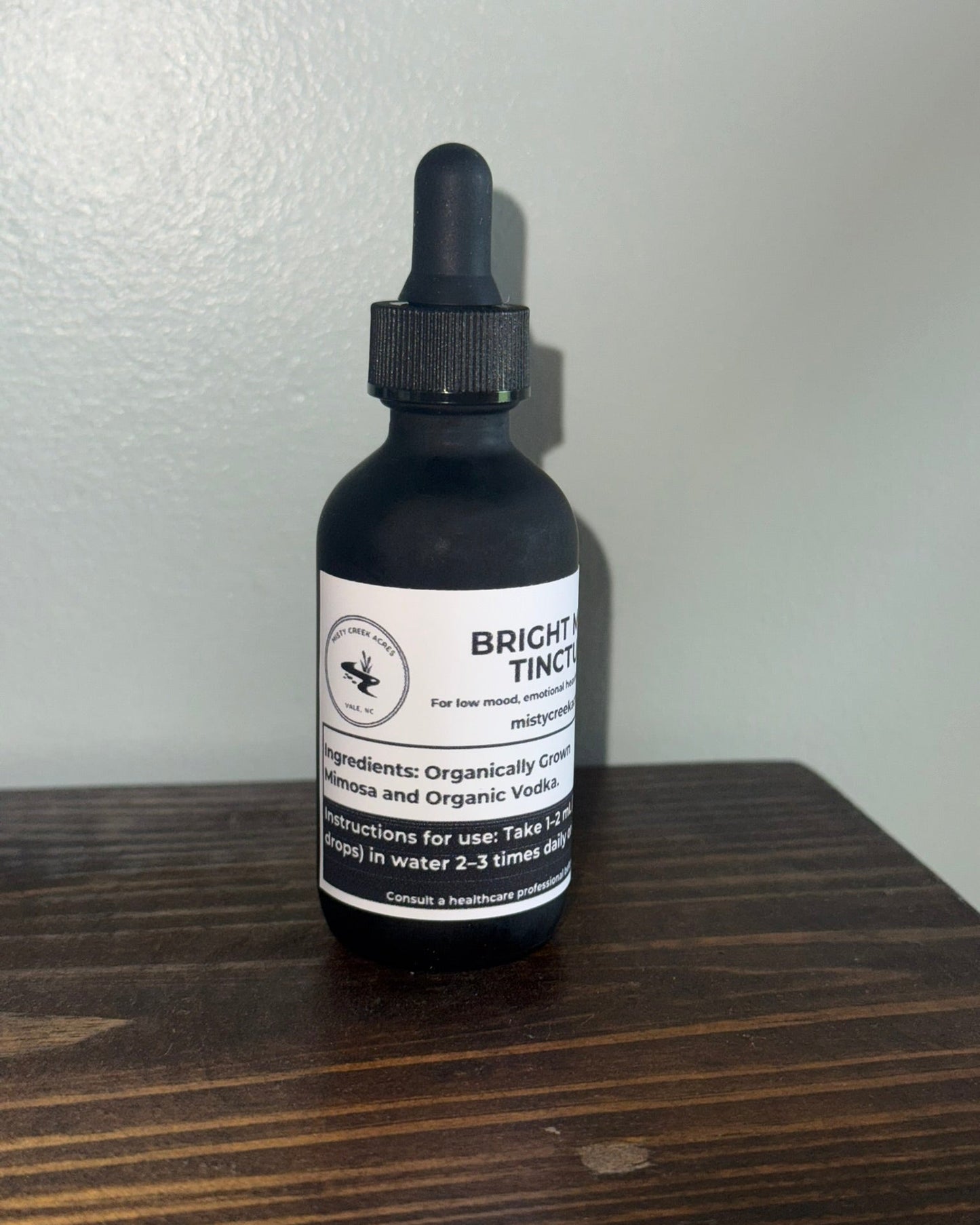 A dark glass dropper bottle with a black cap and a white label reading 'Bright Tincture' and 'miroysses', standing on a dark wooden surface against a light gray wall.