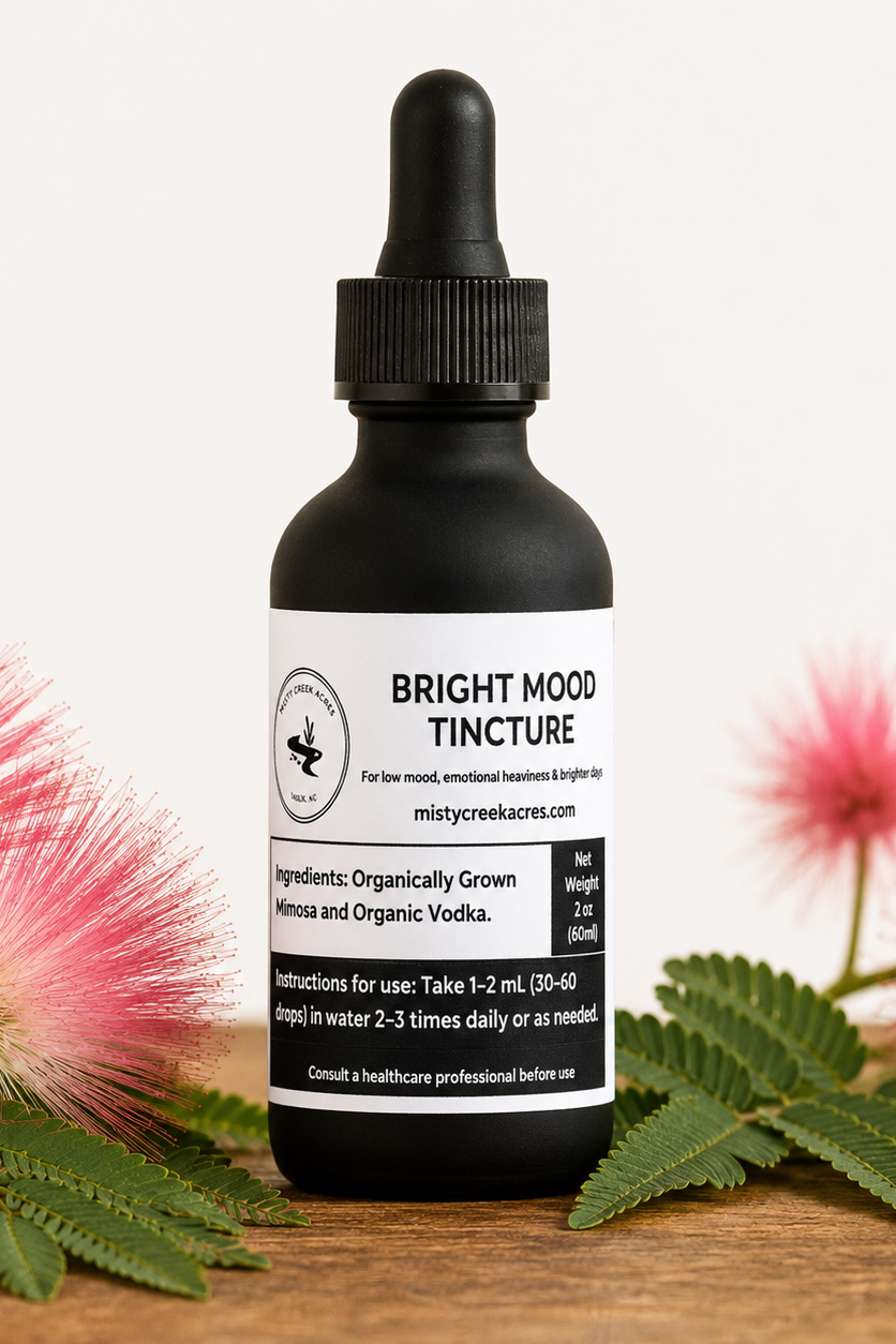 A black dropper bottle labeled 'Bright Mood Tincture' stands on a wooden surface with pink fluffy flowers and green leaves around it. The background is a clean white studio backdrop.