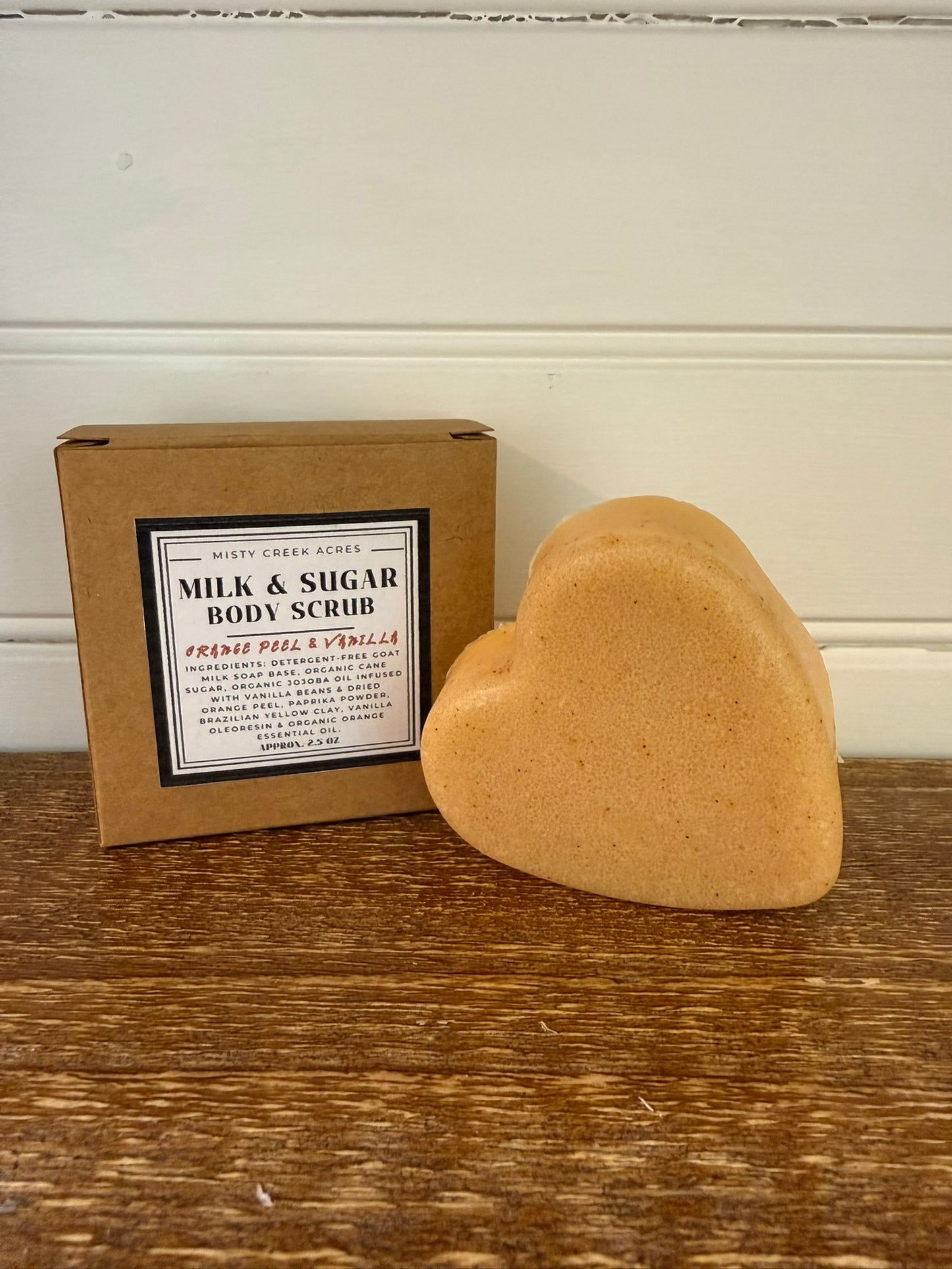 Heart-shaped body scrub next to a cardboard box with product information on a wooden surface.
