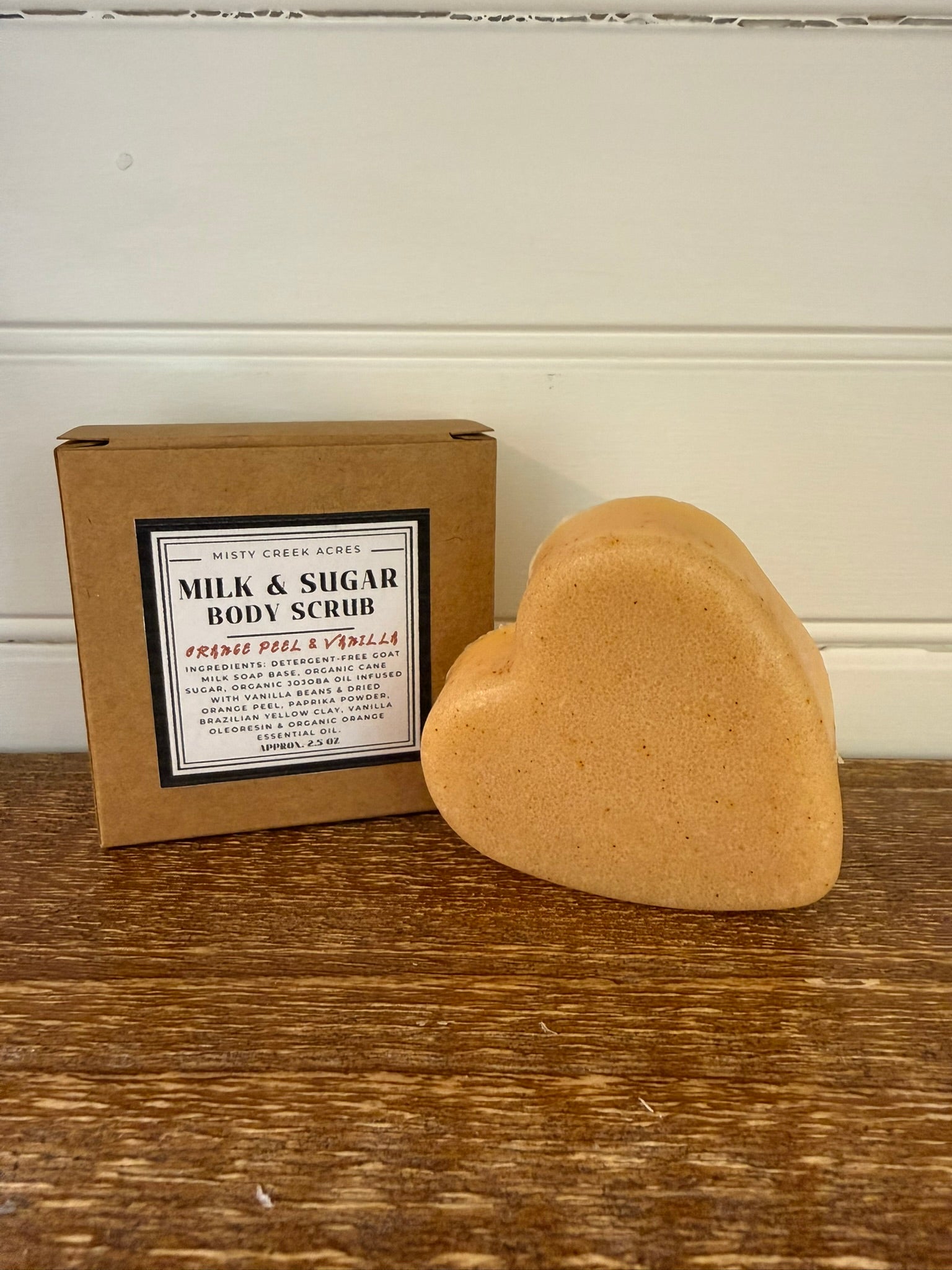 Heart-shaped body scrub next to a cardboard box with product information on a wooden surface.