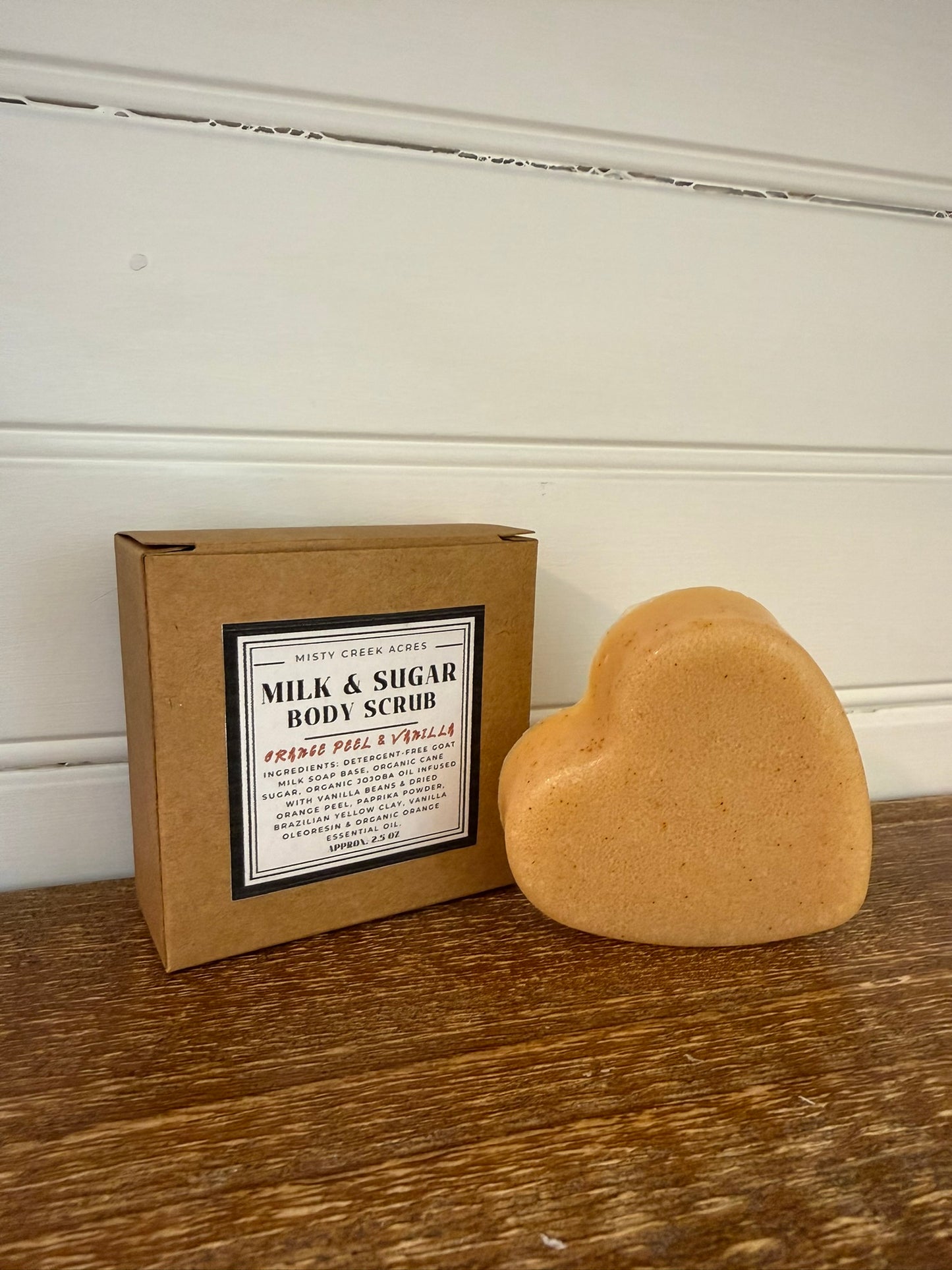 Heart-shaped body scrub next to its packaging on a wooden surface with a white wall background.