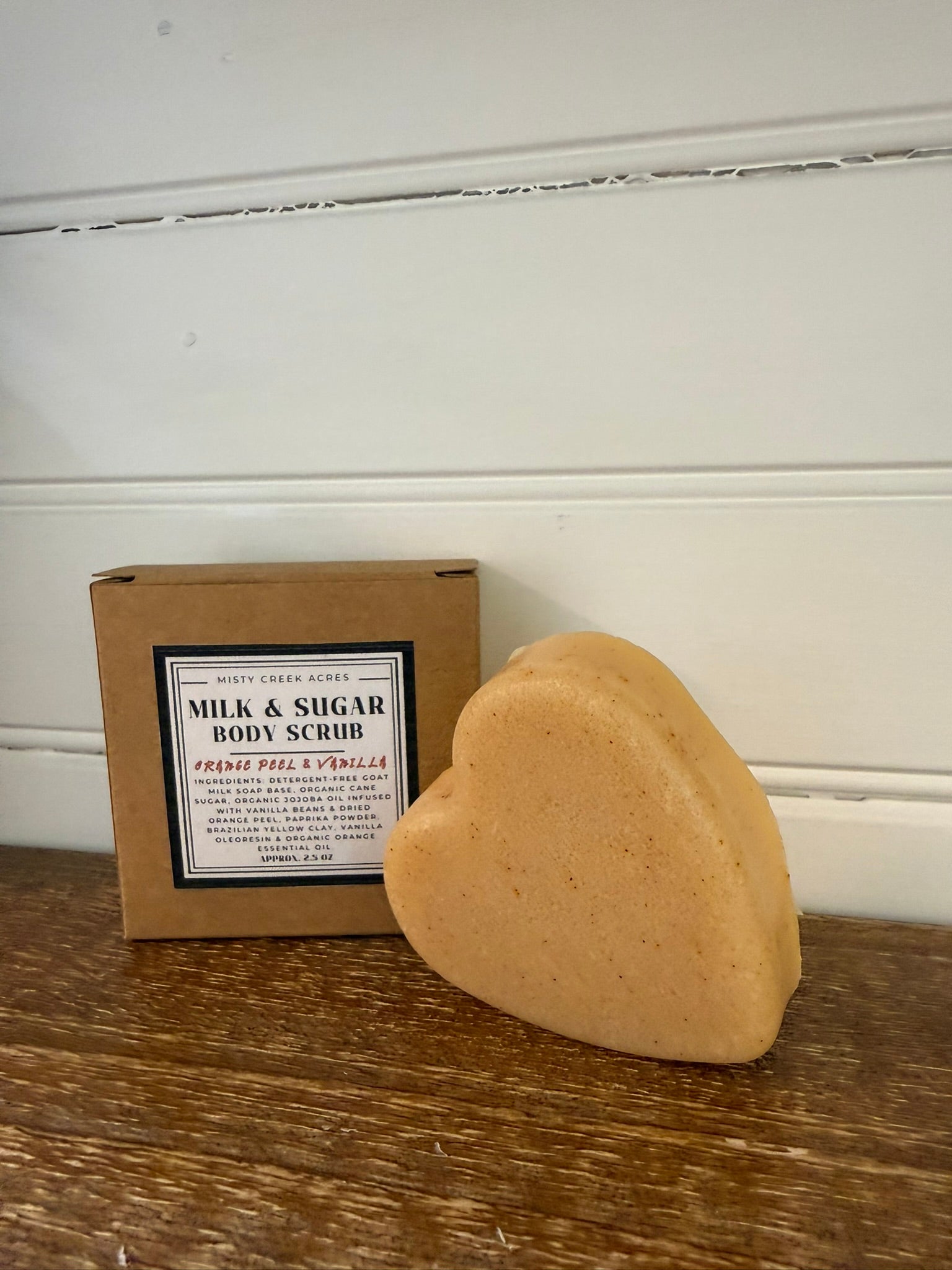 Heart-shaped body scrub bar next to a box labeled 'Milk & Sugar Body Scrub' on a wooden surface.