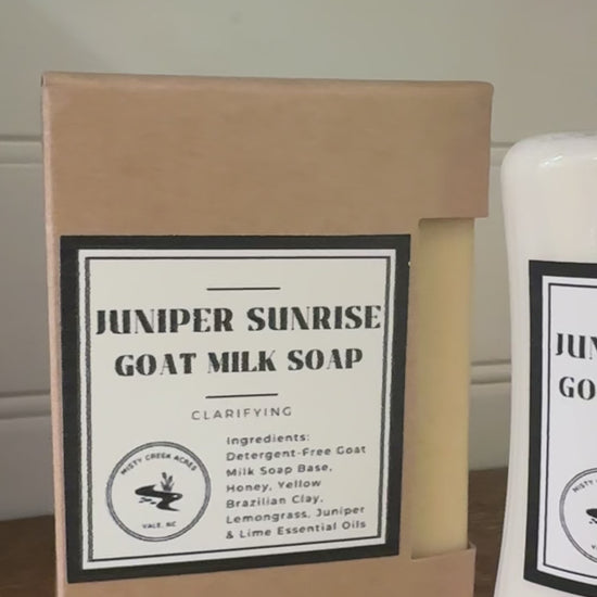 Juniper Sunrise Goat Milk Soap and Goat Milk Lotion set, featuring a kraft paper box of soap and a white bottle of lotion on a wooden surface with a green plant in the background.