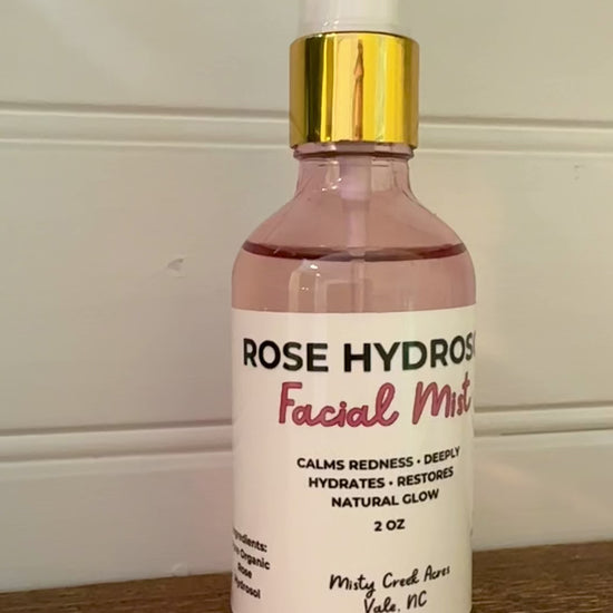 A bottle of Rose Hydrated Facial Toner with pink liquid, a white label, and a gold collar, sitting on a wooden surface against a white paneled wall.