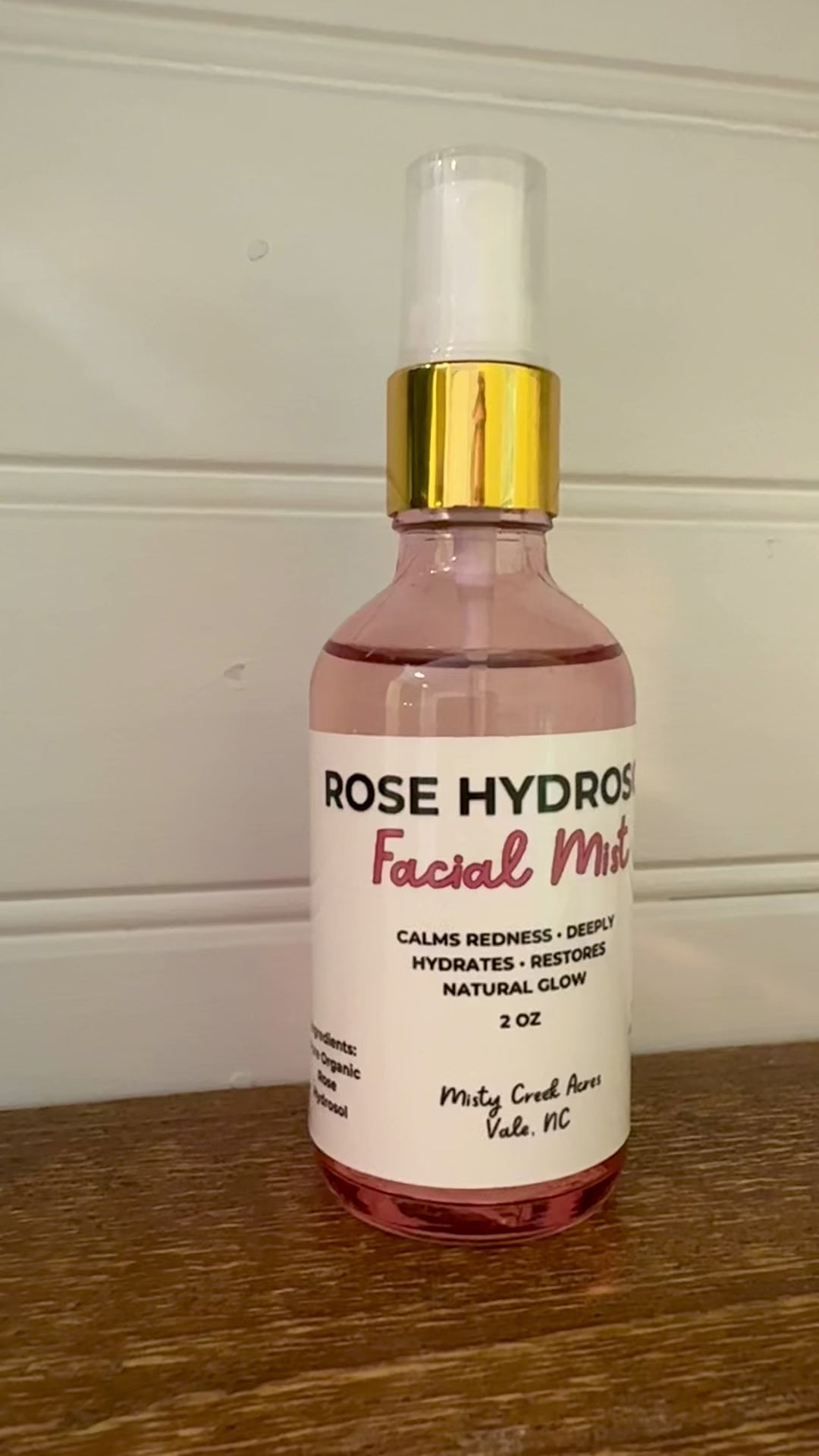 A bottle of Rose Hydrated Facial Toner with pink liquid, a white label, and a gold collar, sitting on a wooden surface against a white paneled wall.