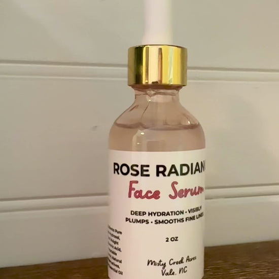 A glass dropper bottle of Rose Rad Face Serum with rose-colored liquid, a white label, and a gold collar with a white dropper cap, sitting on a wooden surface against a white paneled wall.