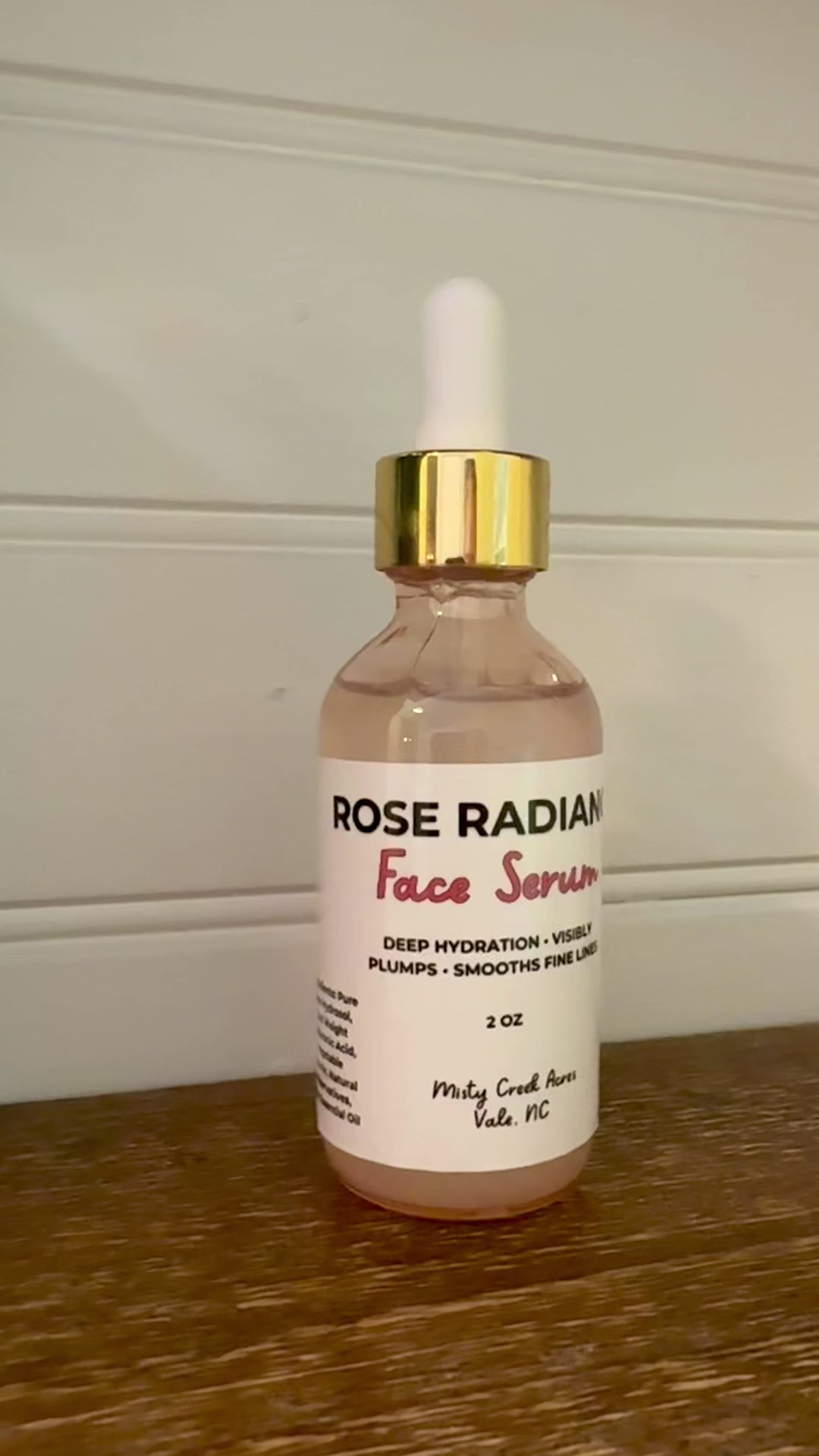 A glass dropper bottle of Rose Rad Face Serum with rose-colored liquid, a white label, and a gold collar with a white dropper cap, sitting on a wooden surface against a white paneled wall.
