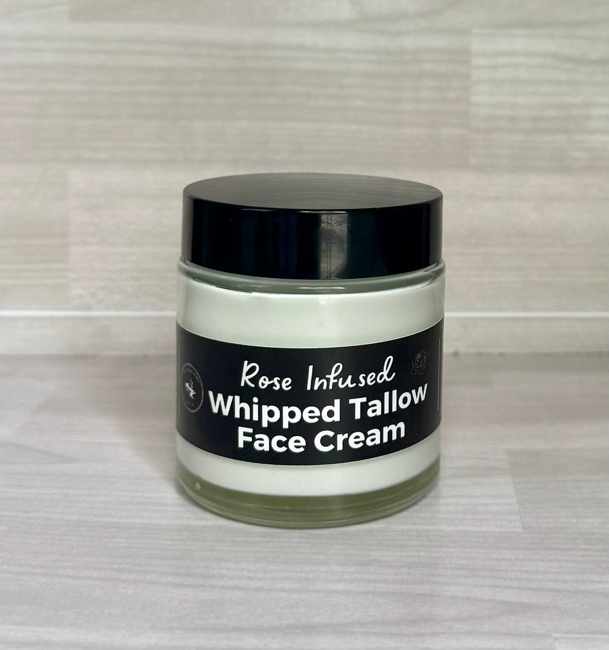 A jar of Rose Infused Whipped Tallow Face Cream with a black lid and black label, set against a light wood grain background.